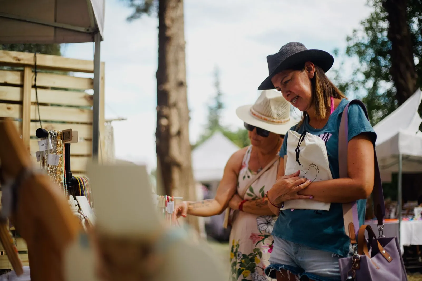 Two women browse handcrafted jewelry at an outdoor market under sunny skies.